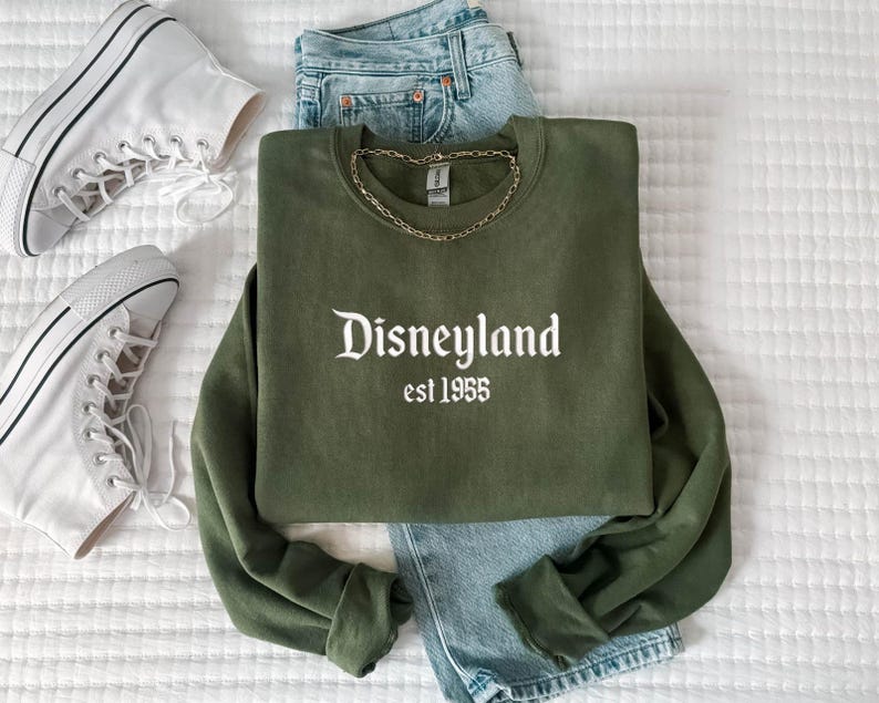 Puede incluir: Una sudadera verde oliva con la palabra "Disneyland" y el a&ntilde;o "est 1955" impresos en blanco. La sudadera se combina con vaqueros de mezclilla azul claro, zapatillas altas blancas y un collar de cadena dorada. Los art&iacute;culos est&aacute;n dispuestos sobre una superficie blanca texturizada.