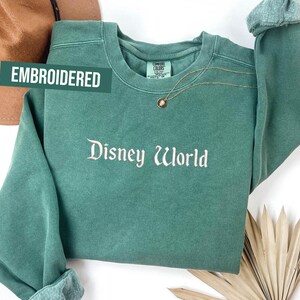 Puede incluir: Una sudadera verde azulado con el texto bordado "Disney World" en escritura blanca. La palabra "EMBROIDERED" est&aacute; impresa en un rect&aacute;ngulo verde. Un collar dorado con un colgante est&aacute; drapeado sobre la sudadera.