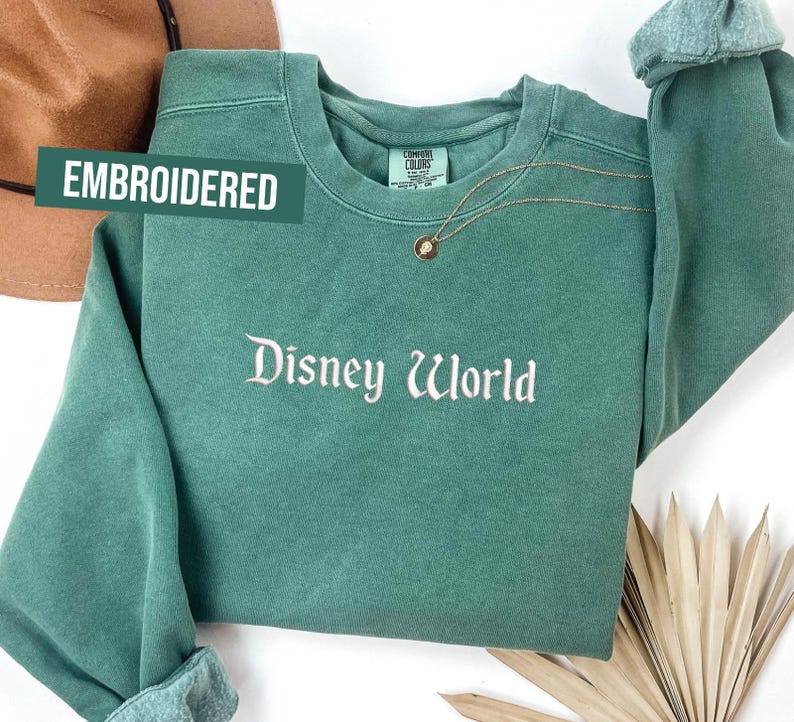 Puede incluir: Una sudadera verde azulado con el texto bordado "Disney World" en escritura blanca. La palabra "EMBROIDERED" est&aacute; impresa en un rect&aacute;ngulo verde. Un collar dorado con un colgante est&aacute; drapeado sobre la sudadera.