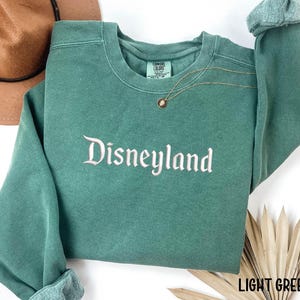 Puede incluir: Sudadera verde claro con la palabra "Disneyland" bordada en escritura blanca. La sudadera tiene mangas largas y cuello redondo. Un collar dorado con un colgante est&aacute; drapeado en la parte delantera. Las palabras "LIGHT GREEN" est&aacute;n en la esquina inferior derecha.