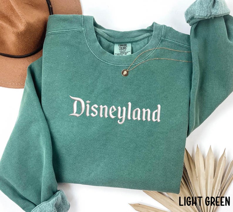 Puede incluir: Sudadera verde claro con la palabra "Disneyland" bordada en escritura blanca. La sudadera tiene mangas largas y cuello redondo. Un collar dorado con un colgante est&aacute; drapeado en la parte delantera. Las palabras "LIGHT GREEN" est&aacute;n en la esquina inferior derecha.