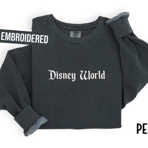 Puede incluir: Sudadera de manga larga color pimienta con las palabras "Disney World" en escritura blanca. La palabra "EMBROIDERED" est&aacute; en una pancarta negra. La palabra "PEPPER" est&aacute; en escritura negra.