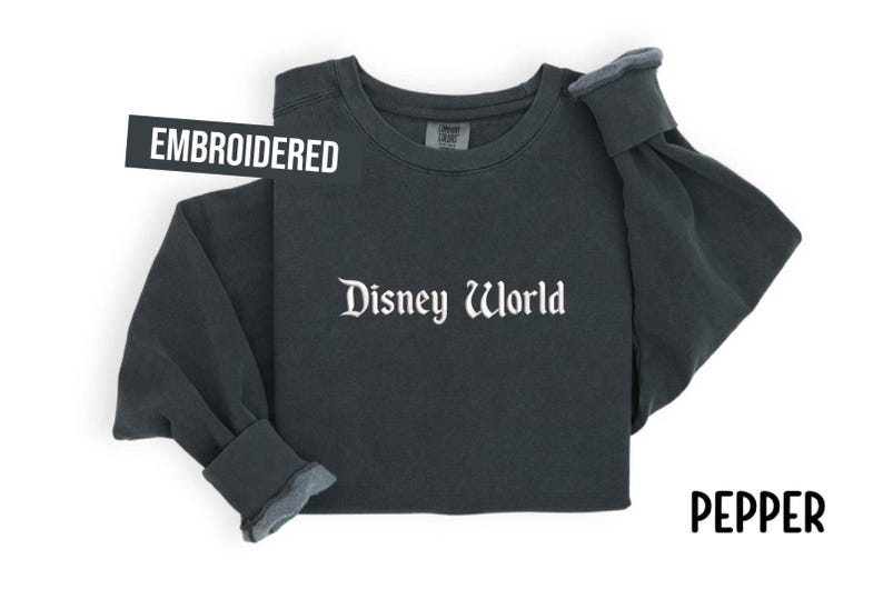Puede incluir: Sudadera de manga larga color pimienta con las palabras "Disney World" en escritura blanca. La palabra "EMBROIDERED" est&aacute; en una pancarta negra. La palabra "PEPPER" est&aacute; en escritura negra.