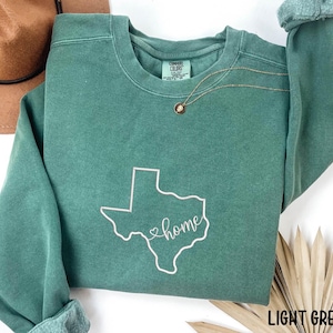 May include: A light green sweatshirt with a white outline of the state of Texas and the word "home" in cursive. The sweatshirt has a crew neck and long sleeves with rolled cuffs. A gold necklace is visible.