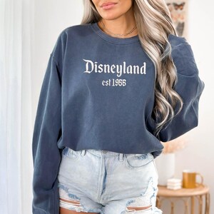 Puede incluir: Una sudadera azul marino con la palabra "Disneyland" en blanco, con una fuente vintage, con "est 1955" debajo. La sudadera es de color azul liso y tiene mangas largas. La persona de la imagen lleva unos shorts vaqueros azules claros.