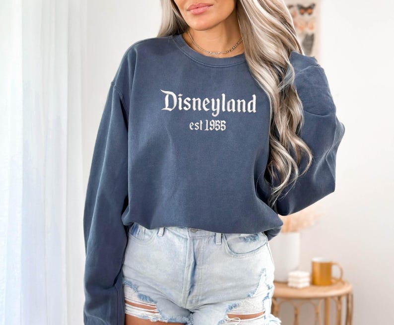 Puede incluir: Una sudadera azul marino con la palabra "Disneyland" en blanco, con una fuente vintage, con "est 1955" debajo. La sudadera es de color azul liso y tiene mangas largas. La persona de la imagen lleva unos shorts vaqueros azules claros.