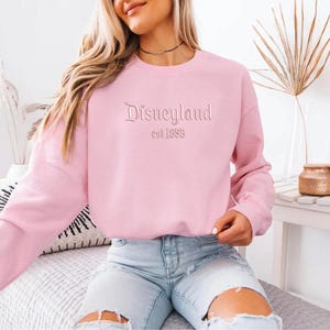 Puede incluir: Sudadera rosa claro con la palabra "Disneyland" y "est 1955" bordada en la parte delantera. La sudadera tiene mangas largas y cuello redondo. La persona de la imagen lleva vaqueros azules claros.