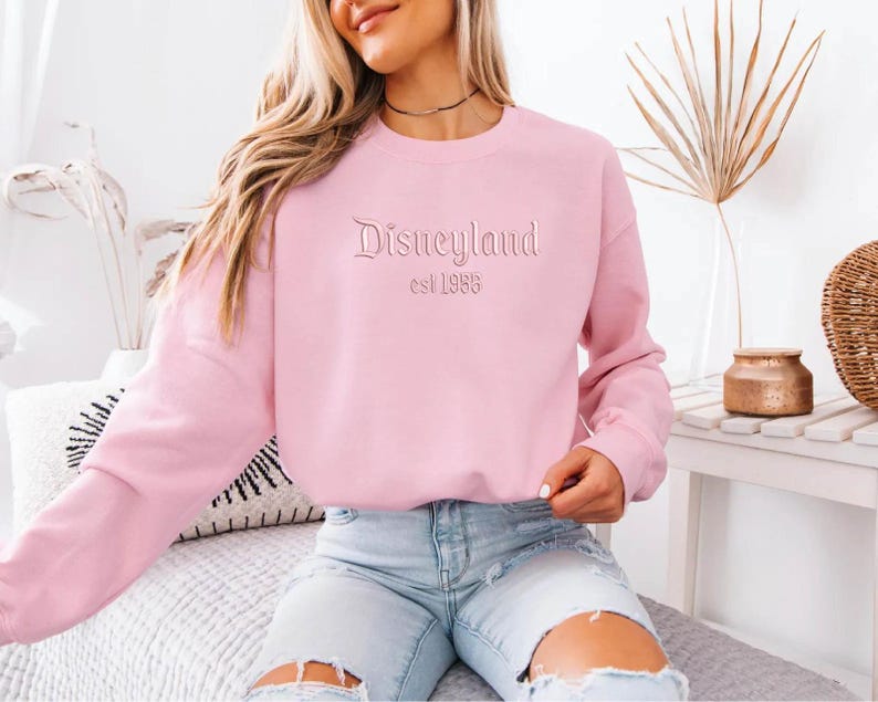 Puede incluir: Sudadera rosa claro con la palabra "Disneyland" y "est 1955" bordada en la parte delantera. La sudadera tiene mangas largas y cuello redondo. La persona de la imagen lleva vaqueros azules claros.