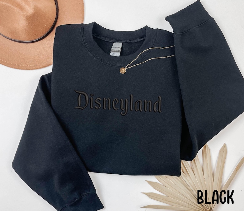 Puede incluir: Sudadera negra con la palabra "Disneyland" bordada en color marr&oacute;n oscuro. Un collar dorado con un colgante est&aacute; drapeado sobre la sudadera. La palabra "BLACK" est&aacute; impresa en blanco en la esquina inferior derecha.