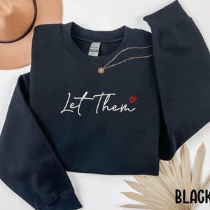 May include: Black sweatshirt with the text "Let Them" in white script with a small red heart above the "m".