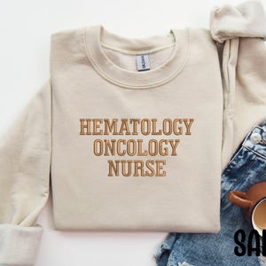 May include: A beige sweatshirt with the text "Hematology Oncology Nurse" in brown lettering.