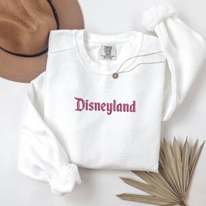 Puede incluir: Una sudadera blanca de cuello redondo con la palabra "Disneyland" bordada en una fuente de escritura. La sudadera tiene mangas largas y una etiqueta en la parte superior del pecho. Un collar dorado con un colgante est&aacute; drapeado en la parte delantera.