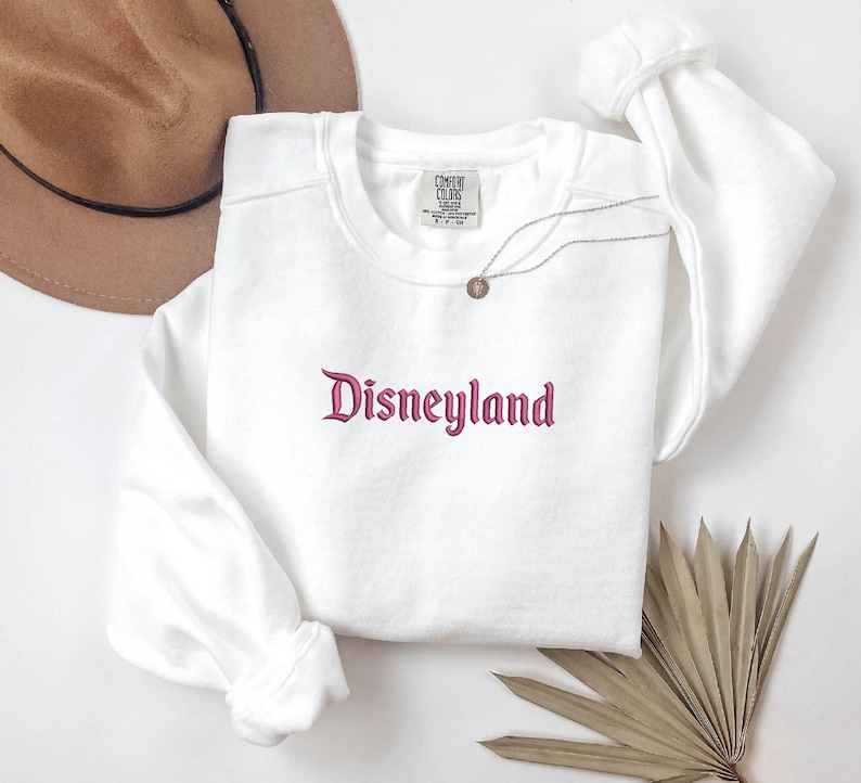 Puede incluir: Una sudadera blanca de cuello redondo con la palabra "Disneyland" bordada en una fuente de escritura. La sudadera tiene mangas largas y una etiqueta en la parte superior del pecho. Un collar dorado con un colgante est&aacute; drapeado en la parte delantera.