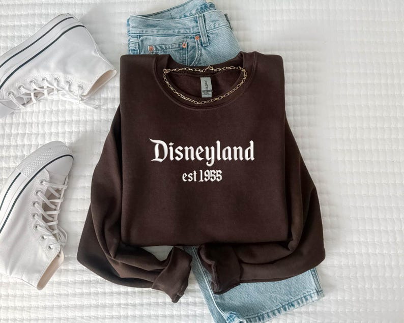 Puede incluir: Una sudadera marr&oacute;n con el texto "Disneyland est 1955" en blanco. La sudadera se combina con vaqueros azul claro y zapatillas de lona blancas. Un collar de cadena dorada est&aacute; drapeado sobre la sudadera.