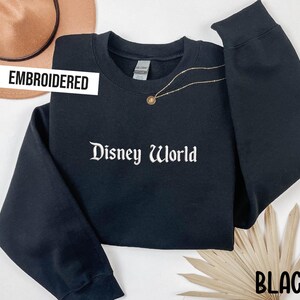 Puede incluir: Sudadera negra bordada con las palabras "Disney World" en escritura blanca. La sudadera tiene mangas largas y cuello redondo. Un collar dorado con un colgante est&aacute; drapeado sobre la sudadera. La palabra "BLACK" est&aacute; en la parte inferior derecha.
