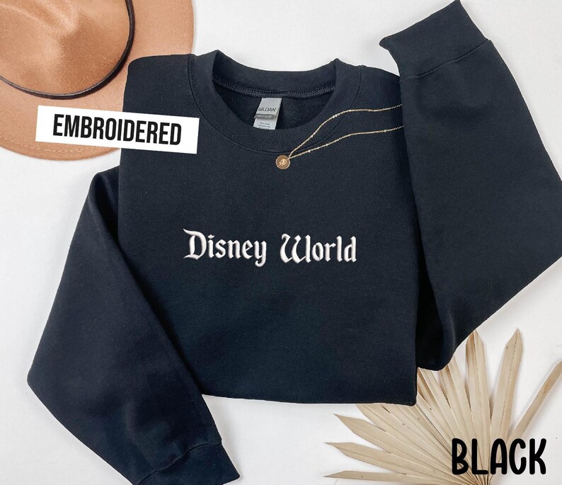 Puede incluir: Sudadera negra bordada con las palabras "Disney World" en escritura blanca. La sudadera tiene mangas largas y cuello redondo. Un collar dorado con un colgante est&aacute; drapeado sobre la sudadera. La palabra "BLACK" est&aacute; en la parte inferior derecha.