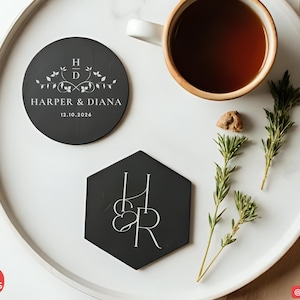 May include: A top-down view of a white tray with a cup of tea, two black coasters, and sprigs of rosemary. One coaster is round with the text "HARPER & DIANA" and the date "12.10.2026". The other coaster is hexagonal with the initials "H & R".