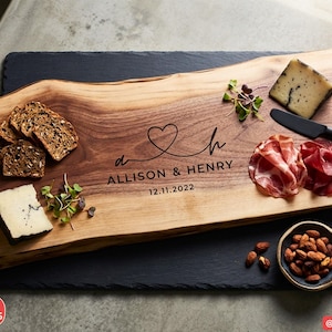 May include: A rectangular live-edge wooden serving board featuring a dark brown wood grain. The board is engraved with a heart design, the letters a and h, the names ALLISON & HENRY, and the date 12.11.2022. It is topped with cheese, meat, and bread.