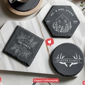 May include: Slate coasters in square, hexagonal, and round shapes, each with a unique design. The coasters feature white text and floral or antler motifs. They are presented on a white marble serving tray.