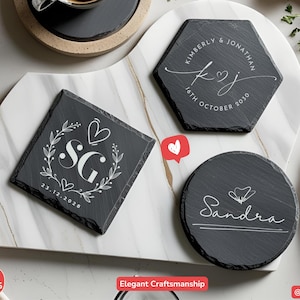 May include: Slate coasters with personalized designs. One square coaster features the initials "SG" with a heart and floral wreath, dated 23.12.2028. A hexagonal coaster displays "Kimberly & Jonathan" and the date 16th October 2030. A round coaster has the name "Sandra".