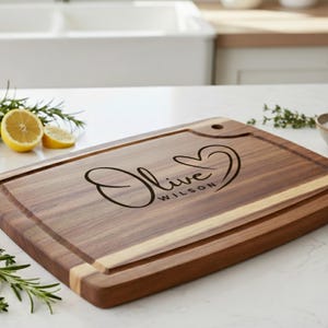 May include: A rectangular wooden cutting board with a juice groove and a hole for hanging. The board features the name "Olive Wilson" in a heart-shaped design. The board is surrounded by lemons, rosemary, and a small bowl of salt.