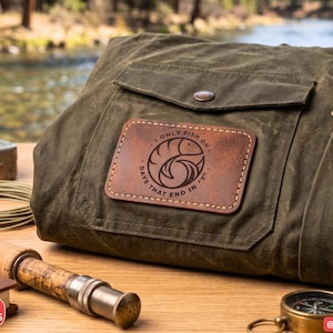 May include: An olive green waxed canvas bag featuring a rectangular brown leather patch with a circular fish graphic and the text I ONLY FISH ON DAYS THAT END IN Y. The bag includes a front flap pocket with a snap button and a side zipper.