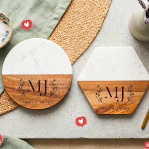 May include: Two personalised coasters, one round and one hexagonal, with a white marble top and a wooden base. The wooden base is engraved with the initials "MJ" and the names "Mino & James". A gold spoon and a small bowl of salt are also visible.