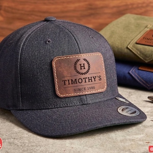 May include: A dark charcoal grey baseball cap with a rectangular brown leather patch on the front. The patch features a laurel wreath icon, the letter H, and the text TIMOTHY'S SINCE 1980. A small circular sticker is visible on the flat brim.