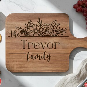 Puede incluir: Tabla de cortar de madera con asa, grabada con las palabras "the Trevor family" y dise&ntilde;o floral. La tabla est&aacute; rodeada de aceitunas, galletas saladas, higos y uvas. La tabla es de color marr&oacute;n claro.