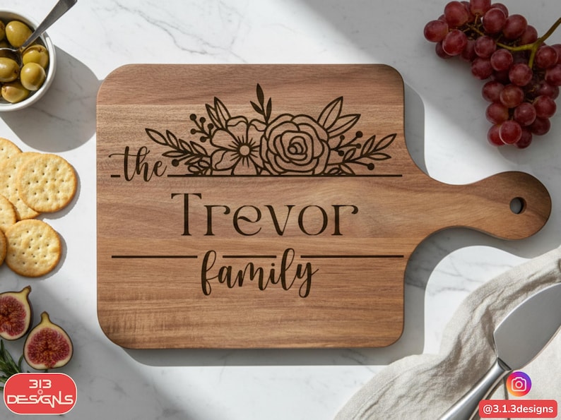 Puede incluir: Tabla de cortar de madera con asa, grabada con las palabras "the Trevor family" y dise&ntilde;o floral. La tabla est&aacute; rodeada de aceitunas, galletas saladas, higos y uvas. La tabla es de color marr&oacute;n claro.