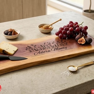 May include: A rectangular wooden serving board with a live edge, featuring a laser-engraved floral design and the text Adora Monty. The board is arranged with a wedge of cheese, a black cheese knife, red grapes, figs, and a small bowl of honey.