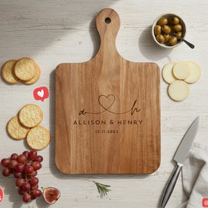 Puede incluir: Tabla de cortar de madera con asa, grabada con un coraz&oacute;n y los nombres "Allison & Henry" y la fecha 12.11.2022. La tabla est&aacute; rodeada de galletas saladas, uvas, aceitunas, rodajas de queso y un cuchillo.