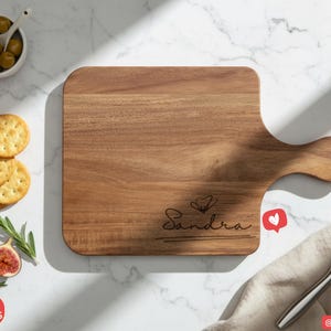 May include: A wooden cutting board with a handle, engraved with the name "Sandra" in cursive. The board is surrounded by olives, grapes, crackers, figs, and a knife. A heart-shaped like button is visible.