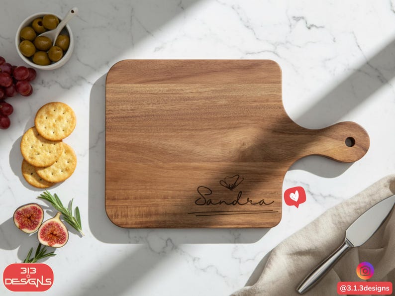 May include: A wooden cutting board with a handle, engraved with the name "Sandra" in cursive. The board is surrounded by olives, grapes, crackers, figs, and a knife. A heart-shaped like button is visible.