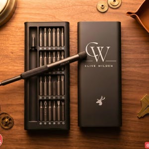 Peut inclure: Un ensemble de tournevis de précision noir dans un boîtier rectangulaire avec un manche assorti. Le boîtier porte le monogramme CW, le nom CLIVE WILSON et une icône de tête de cerf. Un manche de tournevis repose sur le plateau à embouts.