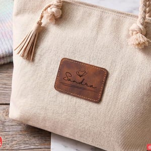 Peut inclure: Un sac fourre-tout en toile beige doté de poignées en corde épaisse avec des extrémités nouées et un pompon décoratif. Un écusson rectangulaire en similicuir marron est centré sur le devant, gravé du prénom Sandra, d'une fleur et d'une ligne.