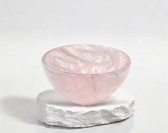 May include: A translucent pink bowl with a marbled appearance, resting on a textured white stone base. The bowl's interior reveals swirling patterns, creating a soft, elegant aesthetic. Ideal for decorative use or as a small serving dish.