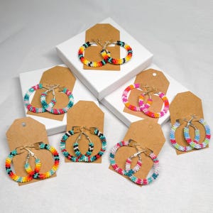 May include: Six pairs of colorful beaded hoop earrings displayed on brown card stock. The earrings are made of small beads in various colors, including pink, blue, green, yellow, orange, and black.