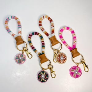 May include: Four beaded keychains with gold-colored hardware. Each keychain features a loop handle with a leather accent and a beaded charm. The beads are in various colors, including pink, white, green, and brown. The charms have a pearl in the center.
