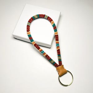 May include: A beaded lanyard with a leather loop and gold-colored ring. The lanyard features a pattern of red, teal, tan, and orange beads. The lanyard is resting on a white surface.