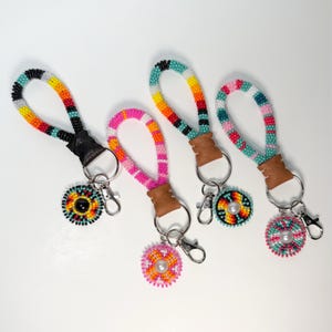 May include: Four beaded keychains with colorful patterns. Each keychain features a loop, a leather accent, a metal ring, and a round beaded charm with a pearl center. The keychains have a variety of colors, including pink, orange, yellow, and turquoise.