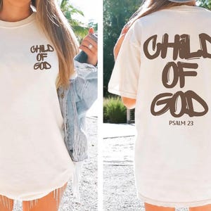 A woman wearing a white t-shirt with the text "CHILD OF GOD" printed on it, standing in a natural outdoor setting.