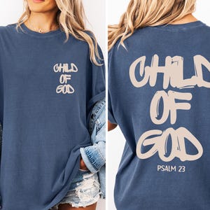 May include: A navy blue oversized t-shirt with the words "CHILD OF GOD" printed in a light beige color on the front and back. The back of the shirt also includes "PSALM 23". The shirt has short sleeves and a relaxed fit.