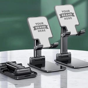 May include: Black adjustable phone stands with the text "YOUR BRAND HERE" on the top. One stand is folded flat, while the other two are upright. The stands are on a white surface.