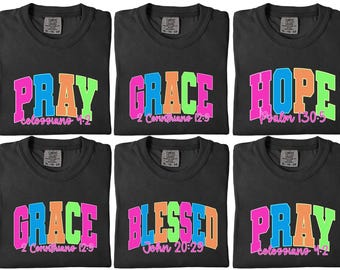 May include: Six black t-shirts with colorful text. Each shirt displays a different word: PRAY, GRACE, HOPE, BLESSED. The words are in a bold, collegiate style font with a neon color scheme. Each shirt also has a Bible verse reference.