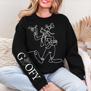K&ouml;nnte beinhalten: Schwarzes Sweatshirt mit einer wei&szlig;en Strichzeichnung von Goofy. Das Wort "GOOFY" ist vertikal auf dem linken &Auml;rmel aufgedruckt. Das Design zeigt Goofy in einer dynamischen Pose, mit einem verspielten Cartoon-Stil.