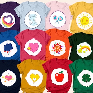 Peut inclure: Plusieurs t-shirts colorés avec des motifs de dessins animés. Chaque t-shirt présente un graphisme différent dans un cercle blanc sur le devant, notamment un cœur arc-en-ciel, une tornade, des sucettes, un soleil souriant, un nuage avec des gouttes de pluie, un cœur avec une étoile, un soleil éclatant, un arc-en-ciel avec des étoiles, une planète avec un cœur, un cœur ampoule, une pomme et un trèfle à quatre feuilles.