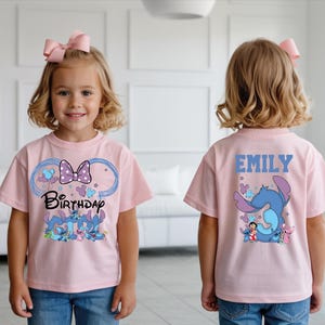 A young girl wearing a pink t-shirt with a cartoon character and the text "Birthday" printed on it, standing in a room with a white wall and a couch.