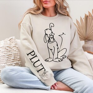 K&ouml;nnte beinhalten: Beiges Sweatshirt mit einer schwarzen Strichzeichnung von Pluto, einem Disney-Cartoon-Hund. Das Wort "PLUTO" ist vertikal auf dem linken &Auml;rmel aufgedruckt. Eine kleine Pluto-Illustration befindet sich ebenfalls auf dem &Auml;rmel. Das Sweatshirt hat eine lockere Passform.