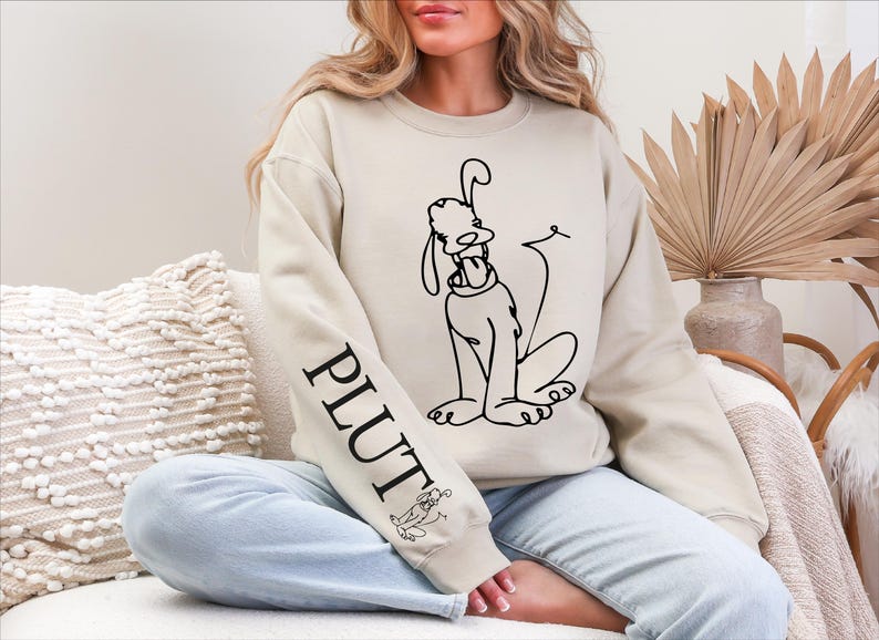 K&ouml;nnte beinhalten: Beiges Sweatshirt mit einer schwarzen Strichzeichnung von Pluto, einem Disney-Cartoon-Hund. Das Wort "PLUTO" ist vertikal auf dem linken &Auml;rmel aufgedruckt. Eine kleine Pluto-Illustration befindet sich ebenfalls auf dem &Auml;rmel. Das Sweatshirt hat eine lockere Passform.